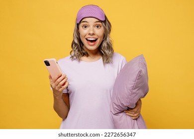 Young fun woman wears purple pyjamas jam sleep eye mask rest relax at home hold pillow use mobile cell phone chatting isolated on plain yellow background studio portrait. Good mood night nap concept