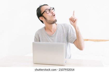 Young Funny Asian Man Looking Up And Pointing Finger Up To Present An Empty Space While Using Laptop Computer In Room. Korean Or Chinese Man Having A Good Time With Funny Jokes.