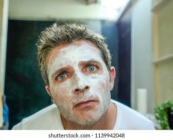 Young Funny Attractive Man At Home Bathroom Looking At Toilet Mirror Applying Facemask In His Face Finding Himself Weird And Ugly With The White Mask In Male Beauty Skin Care And Hygiene Concept