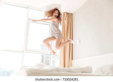 Young funny caucasian woman in pajamas jumping on bed with pillow and smiling at home