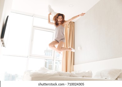 Young funny caucasian woman in pajamas jumping on bed with pillow and smiling at home