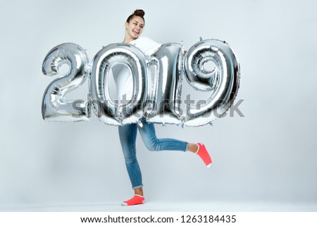 Young funny girl dressed in white t-shirt, jeans and pink socks holding balloons in the shape of numbers 2019 on the white background in the studio