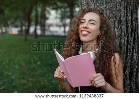 young funny girl student holding a book and sitting under a tree in summer park.