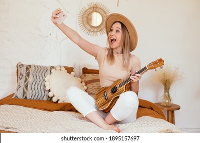 Young Funny Hipster Girl Having Fun And Playing On Small Ukulele Guitar, Singing And Dancing. Wearing A Hat, Joy, Positive Mood. Instagram Live Concert. Making Selfie