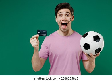Young funny man fan wears basic pink t-shirt cheer up support football sport team hold in hand credit bank card soccer ball watch tv live stream scream isolated on dark green color background studio