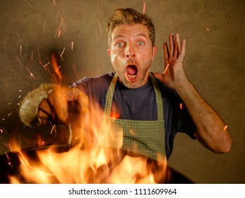 Young Funny And Messy Home Cook Man With Apron In Shock Holding Pan In Fire Burning The Food In Kitchen Disaster And Unskilled Unexperienced Terrible Home Cook At Domestic Cooking Mess