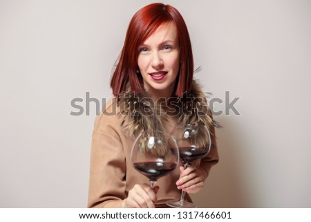 Young funny redhaired woman sommelier with fur cape on white background smile, hold two glasses red wine. Concept alcoholism, addiction, bad habit, collection great rare wines, winelover, expert