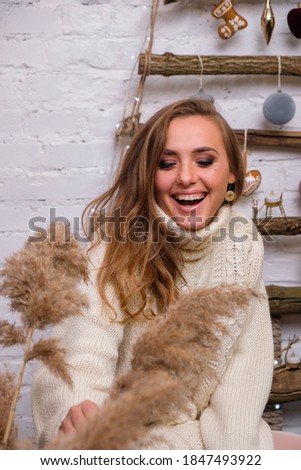 Young, funny woman at home. Minimalist christmas cozy decor on the background. Merry Christmas and Happy Holidays. Blonde, cute, woman in white sweater. New Year's Eve mood.