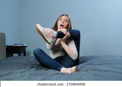 Young Funny Woman Trying To Make Pose Of Yoga, Humor, Laughter. Girl Sitting At Home On The Bed Does Stretching