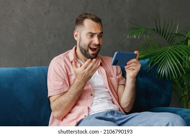 Young Gambling Confused Man In Casual Clothes Using Play Racing App On Mobile Cell Phone Gadget Smartphone For Pc Video Games Sitting On Blue Sofa At Home Flat Indoors Rest Relax On Weekends Free Time