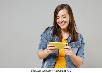 Young Gambling Woman Wear In Denim Jacket Yellow T-shirt Using Play Racing On Mobile Cell Phone Hold Gadget Smartphone For Pc Video Games Isolated On Grey Background Studio. People Lifestyle Concept