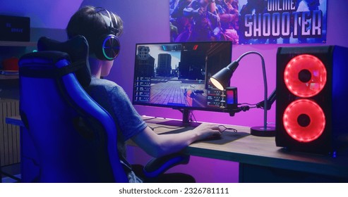 Young gamer in headphones plays in first person shooter on computer. Online video game live streaming or esports tournament. Desk illuminated by RGB LED strip light. Gaming at home concept. Back view.