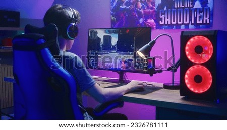 Young gamer in headphones plays in first person shooter on computer. Online video game live streaming or esports tournament. Desk illuminated by RGB LED strip light. Gaming at home concept. Back view.