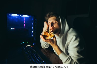 Young gamer sits at home at the table at the computer and eats pizza. Young man eating pizza at break after computer game. Night gaming with fast food man having pizza and using computer - Powered by Shutterstock