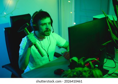 A Young Gamer Spends Time At The PC In The Evening. A Streamer Guy With Headphones Plays Computer Games And Gets Angry. A Man With A Hammer In His Hands In A Rage Wants To Hit The Screen