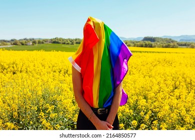 Young Gay Boy Covers His Face With Rainbow Gay Pride Flag, In Yellow Flowers Field At Spring Sunny Day. LGBT, Homosexual Discrimination Concept.