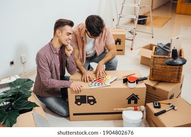 Young Gay Couple Sitting On The Floor Of A New Apartment They Moved In, Looking At Color Samples To Choose The Right Color For Their Wall, While Renovating Apartment. Moving Into A New Home.