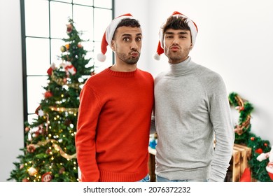 Young Gay Couple Standing By Christmas Tree Wearing Hat Looking At The Camera Blowing A Kiss On Air Being Lovely And Sexy. Love Expression. 