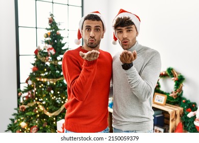 Young Gay Couple Standing By Christmas Tree Wearing Hat Looking At The Camera Blowing A Kiss With Hand On Air Being Lovely And Sexy. Love Expression. 