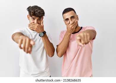 Young Gay Couple Standing Over Isolated Background Laughing At You, Pointing Finger To The Camera With Hand Over Mouth, Shame Expression 