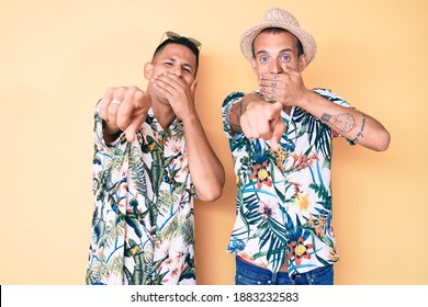 Young Gay Couple Of Two Men Wearing Summer Hat And Hawaiian Shirt Laughing At You, Pointing Finger To The Camera With Hand Over Mouth, Shame Expression 