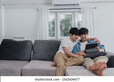 Young Gay Couple Using Tablet At Home. Asian LGBTQ+ Men Happy Relax Fun Using Technology Watching Movie In Internet Together While Lying Sofa In Living Room Concept.