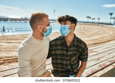 Young Gay Couple Wearing Medical Mask Sitting On The Bench At The Beach.