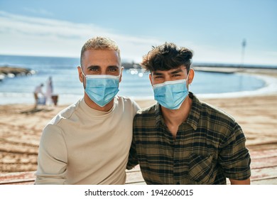 Young Gay Couple Wearing Medical Mask Sitting On The Bench At The Beach.