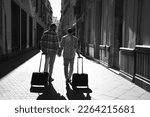 A young gay male couple walks down a street with their suitcases. The couple goes on a trip. The photo is taken from behind and in black and white. Vacation and travel concept.