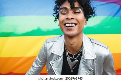 Young gay man embracing his queer identity. Happy gay man smiling cheerfully while standing against a pride flag. Gender non-conforming young man wearing glitter eyeshadow against the LGBTQ+ flag. - Shutterstock ID 2002961945