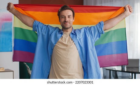 Young Gay Man Looking At Camera Holding Rainbow Flag. Homosexual Guy In Lgbt Flag Smile At Camera Standing At Home