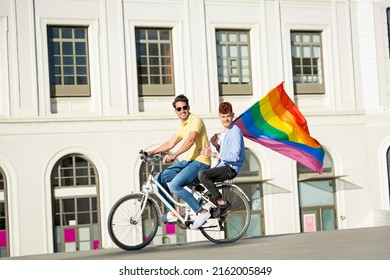 Young Gay Men Couple Riding Bicycle Holding Gay Pride Flag In City. Lgbt Concept