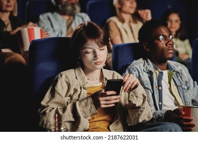 Young Gen Z Caucasian Woman Getting Bored With Movie Scrolling News Feed In Social Network On Smartphone At Cinema