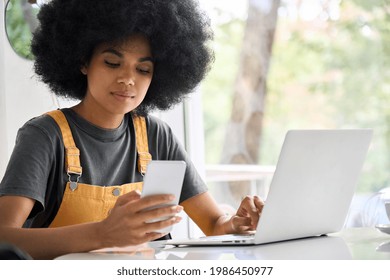 Young Generation Z Black College Student Girl With Afro Hair Sitting At Table In Cafe Using Mobile Shop Marketing Application Online With Computer Working Texting Surfing Internet.