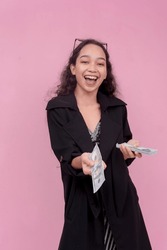 A Young And Generous Asian Woman Gives A Hundred Dollar Bill Tip From Her Wad Of Cash. An Ostentatious Lifestyle. Isolated On A Pink Background.