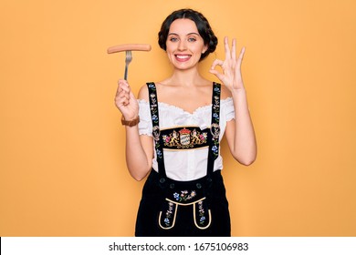 Young German Woman With Blue Eyes Wearing Octoberfest Dress Holding Fork With Sausage Doing Ok Sign With Fingers, Excellent Symbol