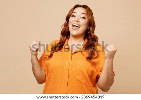 Young ginger chubby overweight woman wear orange shirt casual clothes doing winner gesture celebrate clenching fists say yes isolated on plain pastel light beige background studio. Lifestyle concept