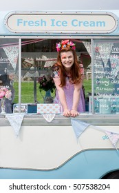 Young ginger haired lady with facepaint smiling wearing vintage dress and flower headband stood in ice cream van. Portrait image with text space