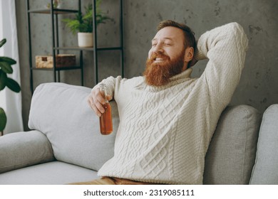 Young ginger man wear casual clothes drink ale hold hands behind neck close eyes sits on grey sofa couch stay at home hotel flat rest relax spend free spare time in living room indoor. Lounge concept - Powered by Shutterstock