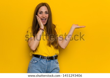 Young ginger redhead woman holds copy space on a palm, keep hand over cheek. Amazed and delighted.