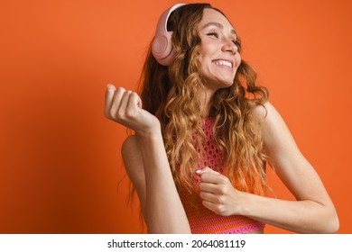 Young Ginger Woman Dancing While Listening Music With Headphones Isolated Over Orange Wall