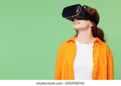 Young ginger woman she wears orange shirt white t-shirt casual clothes watching in vr headset pc gadget look aside isolated on plain pastel light green background studio portrait. Lifestyle concept - Powered by Shutterstock