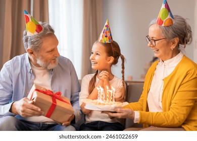 A young girl, adorned with a festive party hat, beams with joy as her grandparents, also wearing colorful party hats, present her with a birthday cake and a wrapped gift during an indoor celebration - Powered by Shutterstock