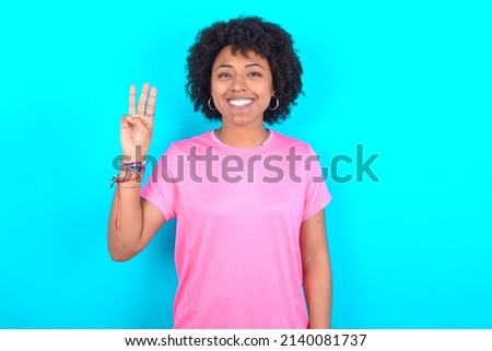 young girl with afro hair style wearing sport pink t-shirt over blue background showing and pointing up with fingers number three while smiling confident and happy.
