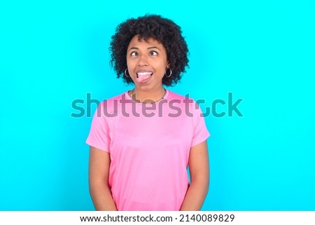 young girl with afro hair style wearing sport pink t-shirt over blue background showing grimace face crossing eyes and showing tongue. Being funny and crazy