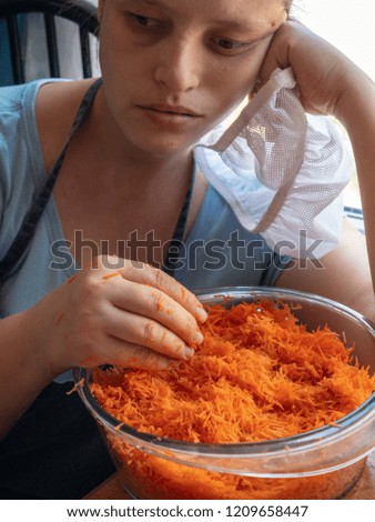 Young girl in apron and a cook's cap prepares fresh carrot salad in kitchen. Woman cook rubs carrots through grater, adds garlic, salt and mayonnaise