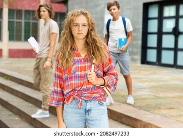 Young Girl With Backpack Standing Near Entrance To School Building. Teenagers Leaving School In Background..