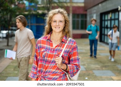 Young Girl With Backpack Standing Near Entrance To School Building, Looking In Camera And Smiling. Teenagers Leaving School In Background.