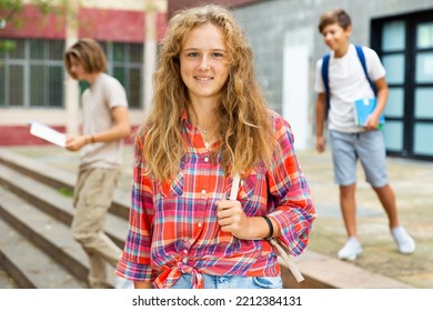 Young Girl With Backpack Standing Near Entrance To School Building, Looking In Camera And Smiling. Teenagers Leaving School In Background.