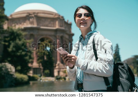 young girl backpacker reading on line guidebook on cellphone visiting Palace of Fine Arts San Francisco California. asian woman in sungalsses holding smart phone smiling looking at blue sky.